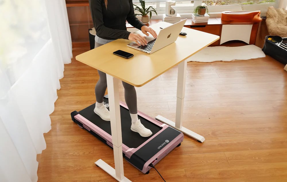 A person in a black sweatshirt works on a laptop at a wooden desk above a pink treadmill