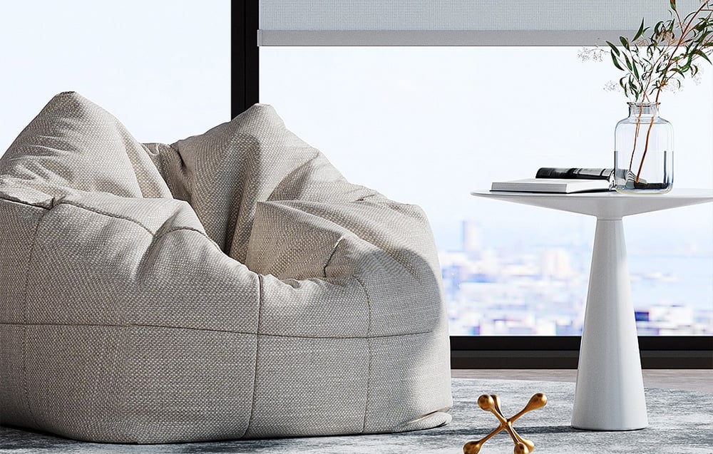 Gray pouf in the living room next to a white side table with decorations by a large window with a blind
