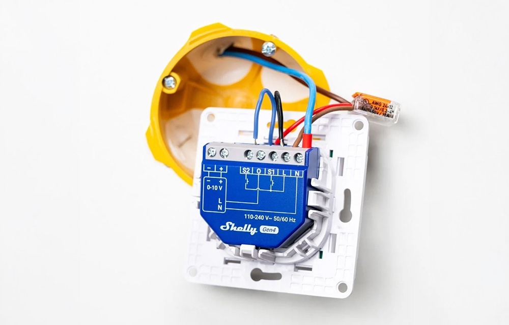 Blue Shelly Gen4 module with screw terminals in a white mounting frame, next to a yellow flush-mount box with visible cables