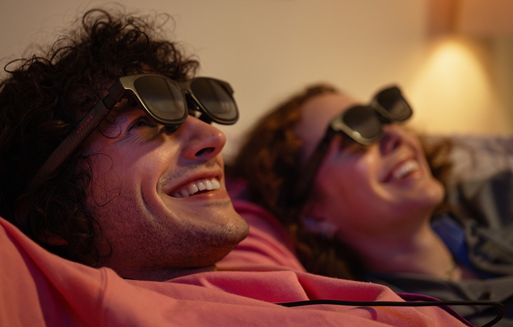 A woman and a man in XR glasses laugh while lying in a room with warm lighting