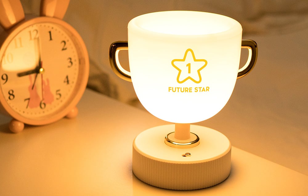 SuperFire W-006 Champion Light children's bedside lamp shining with white light. The device is set on the cabinet next to the alarm clock with rabbit ears.