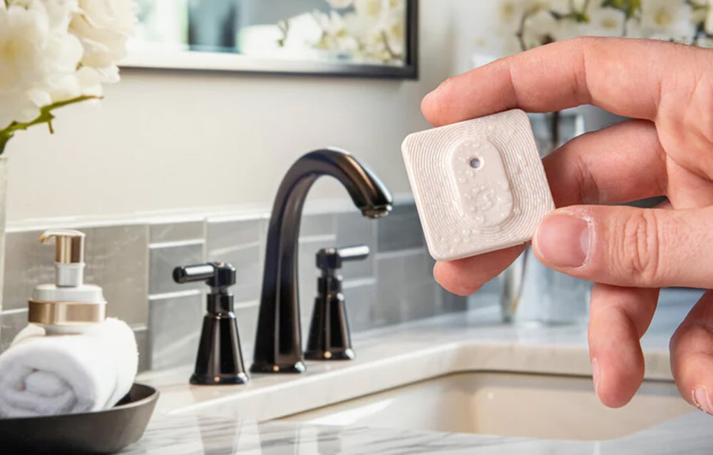 A hand holding a small, wet Shelly H&T; Zigbee sensor against the background of a sink and bathroom faucet.