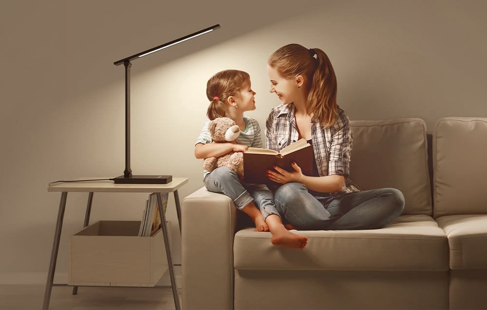 A woman and a child sit on a sofa reading a book next to a lit desk lamp on a side table.