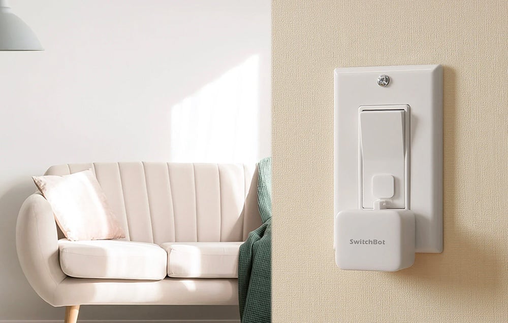 A white SwitchBot switch mounted on a traditional wall light switch in the living room.