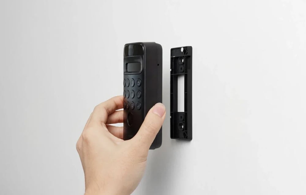 A hand mounting a black SwitchBot numeric keypad on a wall mounting bracket