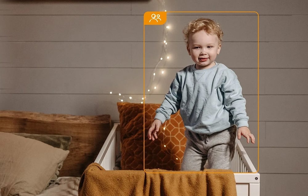 A small child in a blue sweatshirt stands in a crib; the figure is framed by an orange border with an icon of two people