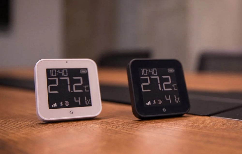 A white and black digital temperature sensor with a hygrometer standing side by side on a wooden desk
