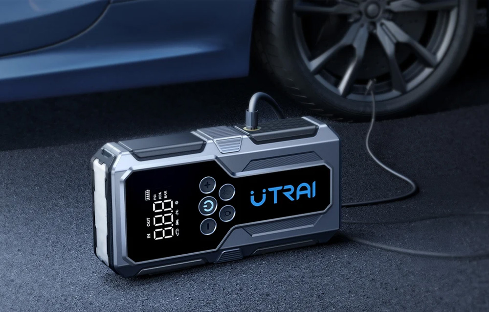 A gray car starter with a display lies on the road and inflates the tire of a blue car using a hose.