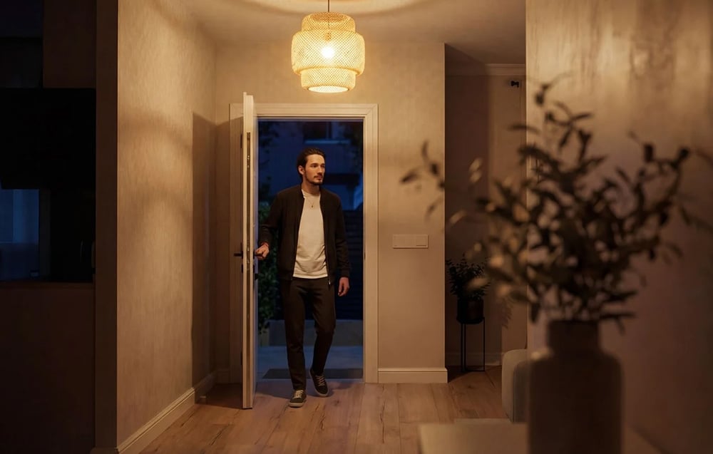 A man in a black jacket and a light-colored T-shirt walks through an open door into a hallway bathed in warm light