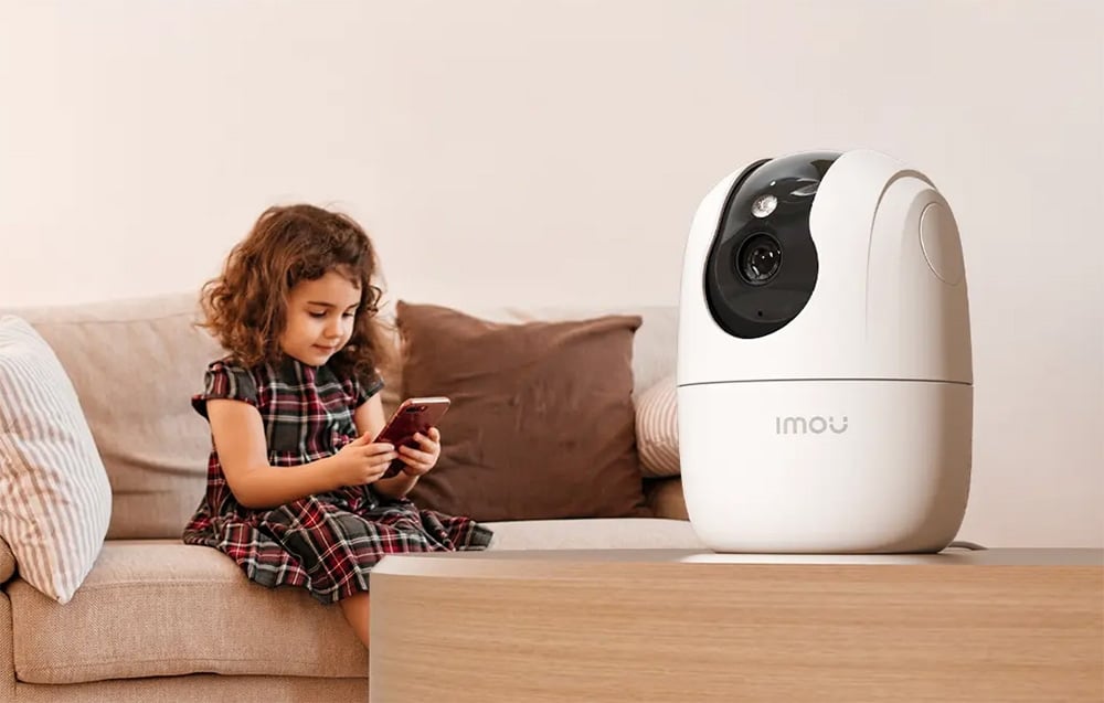 White Imou camera on a table, with a girl sitting on a couch holding a phone in the background