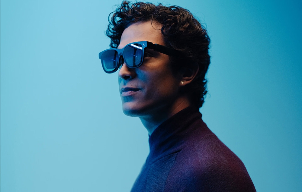 A close-up of the profile of a man with curly hair wearing dark glasses against a light blue background.