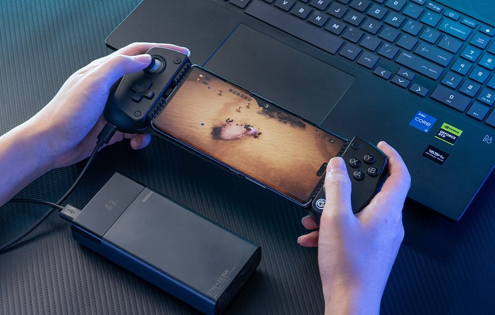 A person playing on a smartphone mounted in a black GameSir controller with a powerbank plugged in next to a laptop.
