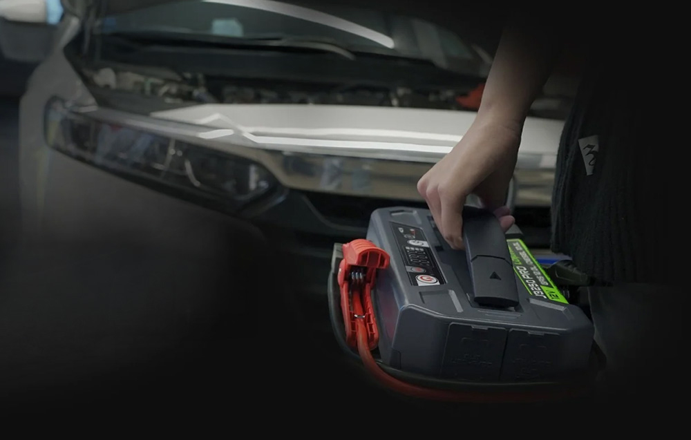 A portable Lokithor jump starter held by the handle with the car hood open in the evening.