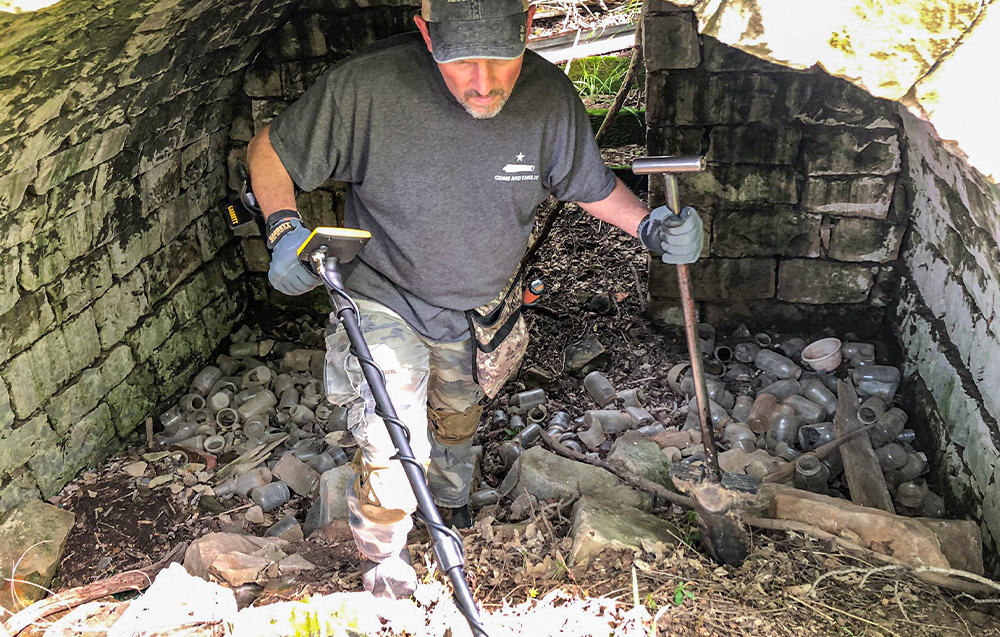 A man with a metal detector searches the inside of a stone cellar full of jars.