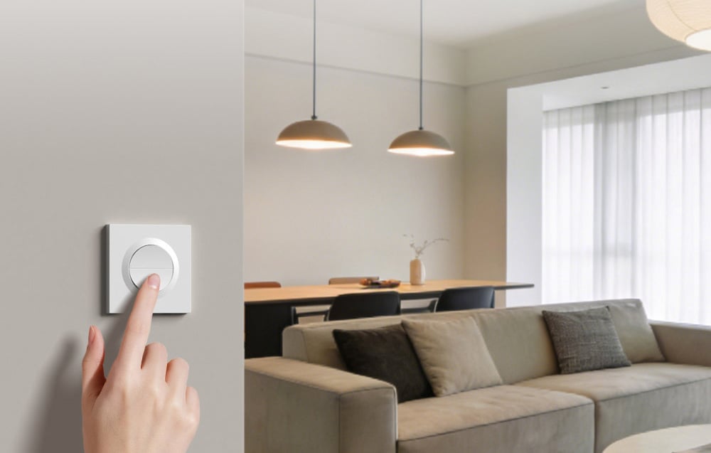 White round SONOFF switch mounted on a bright wall in a modern living room with sofa and dining room in the background