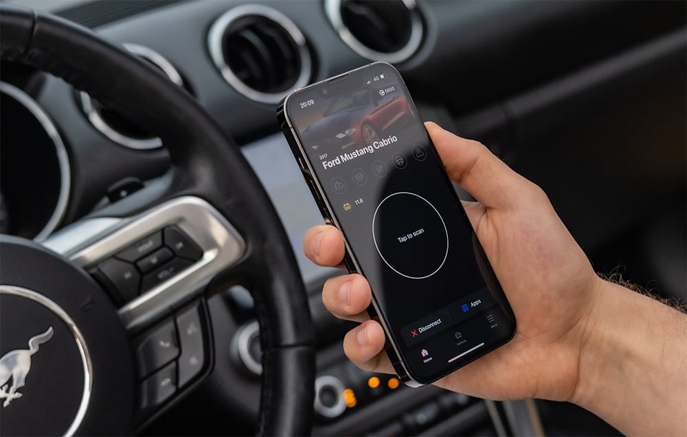 Hand holding smartphone with Carly app displaying Ford Mustang Convertible data inside the car