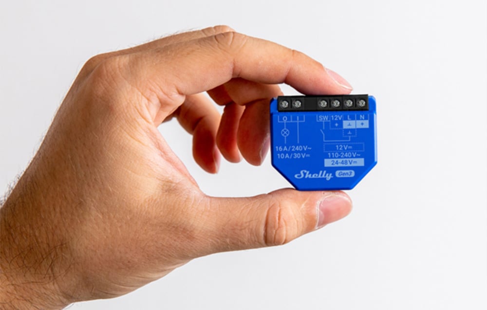 A hand holding a blue Shelly Gen3 module on a white wall background