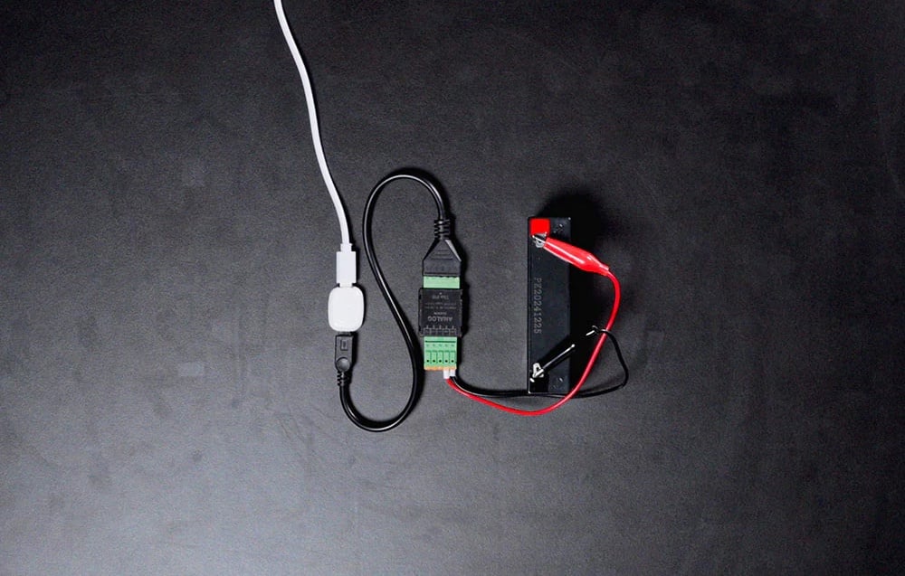 The device connected to the battery with alligator clips on a dark background, with cables and a visible serial number
