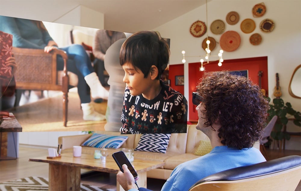 A person sitting in the living room watches an image floating in the air, depicting a boy in a Christmas sweater