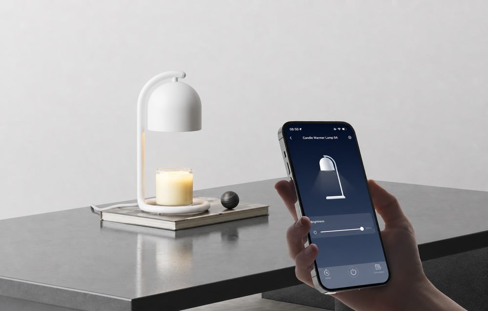 A hand holding a smartphone with the SwitchBot app showing the slider set to 100%, with a white candle warmer and a scented candle in a glass container in the b