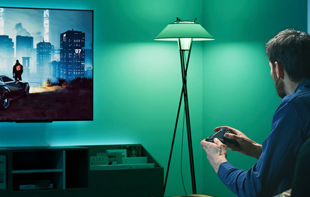 A man holding a controller, sitting in front of the TV in a room lit by turquoise light.