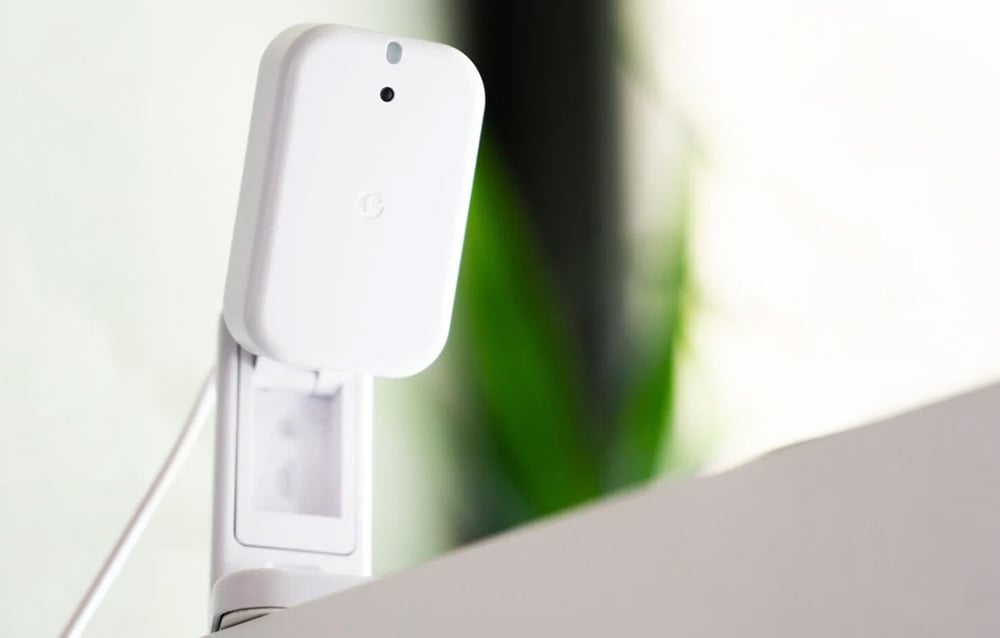 A white electronic device on a desk, mounted on a white clamp, with a cable