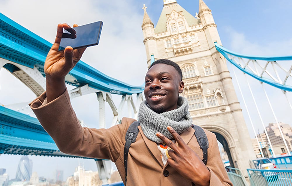 A smiling man taking a selfie with his smartphone against the backdrop of Tower Bridge in London.