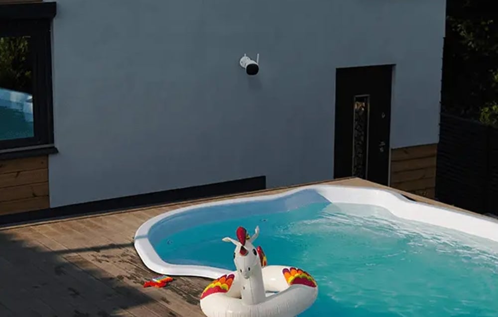 A backyard pool with an inflatable unicorn, with a white building wall in the background featuring a mounted black security camera