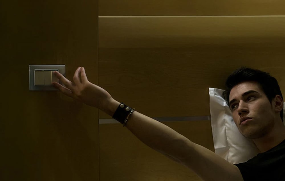 A man lying on a bed reaches toward the wall, pressing a square light switch.