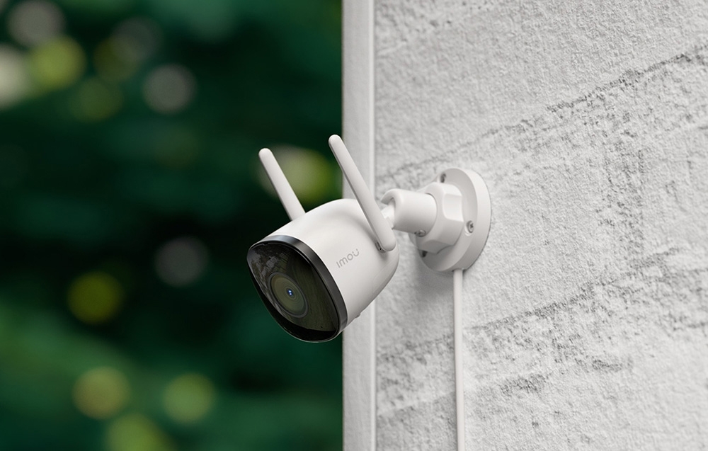 A white Imou outdoor camera with two antennas, mounted on a light-colored, rough-textured building wall
