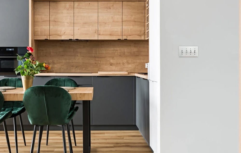 A modern kitchen with wooden and gray cabinet fronts. Green chairs are at the table, and a metal panel with four switches is on the wall.