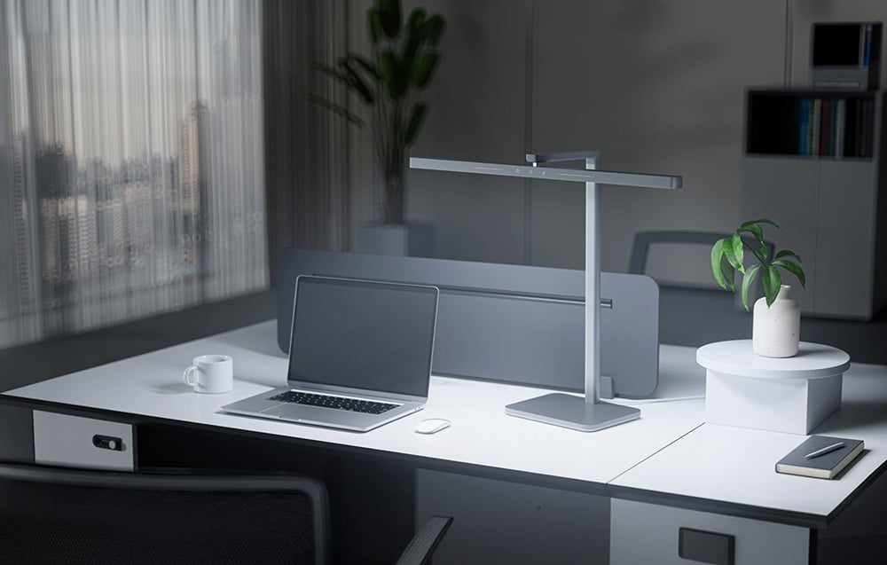A silver, elongated LED desk lamp illuminating a desk with a laptop and a mug in a modern office