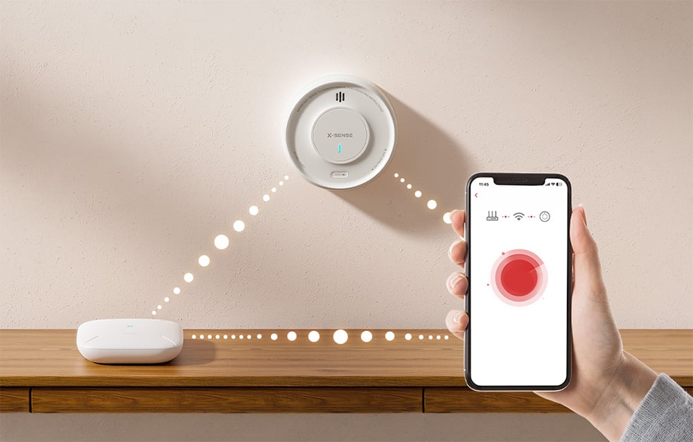 A smartphone displaying an alarm, wirelessly connected to a wall-mounted smoke detector and a countertop base station