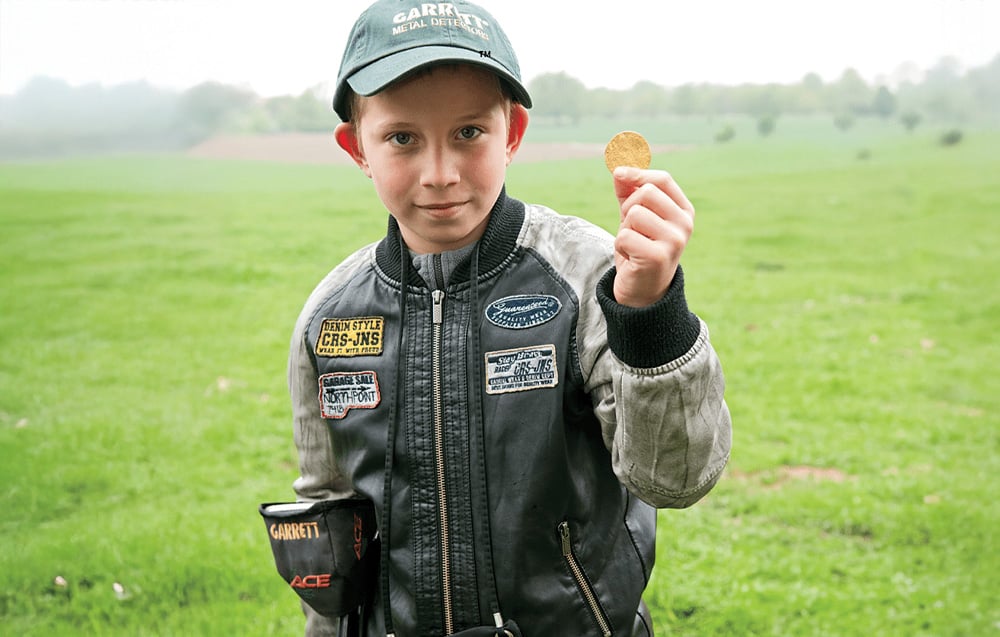 A boy holding a coin and the metal detector against a green field.