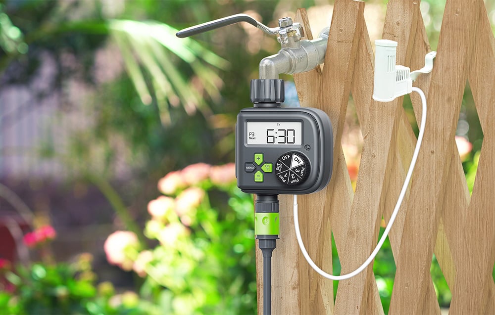 Irrigation controller mounted on a faucet next to a wooden fence, with a hose and rain sensor connected via a white cable