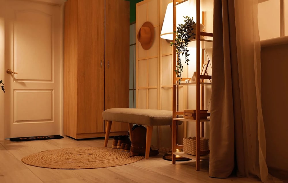A bright, warmly lit entryway with a closet, coat rack, bench, and a floor lamp next to a bookshelf