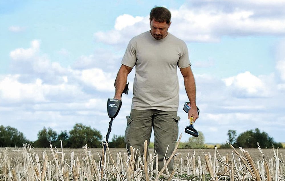 A man walks around a field with a metal detector in one hand and a shovel in the other.