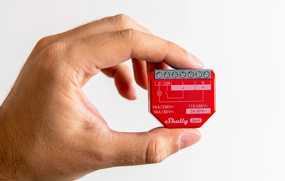 A hand holds a small red Shelly Gen4 controller module with visible connector and technical parameter markings