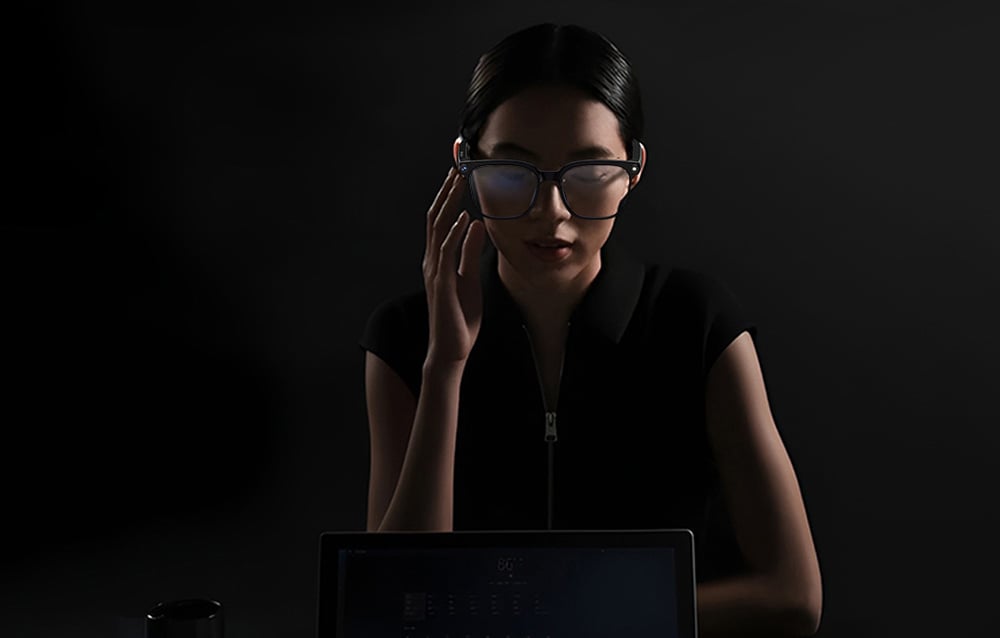 A woman wearing glasses working at a computer in a dark room, touching the temple with her hand.