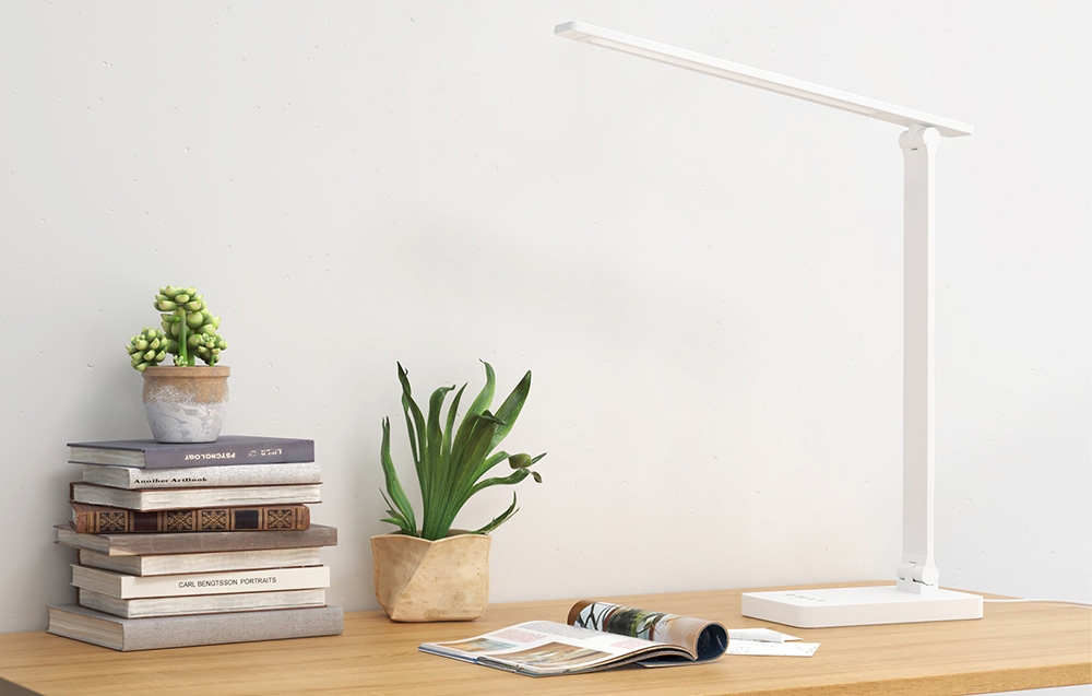 A modern white desk lamp on a wooden table next to a stack of books, potted plants, and an open magazine