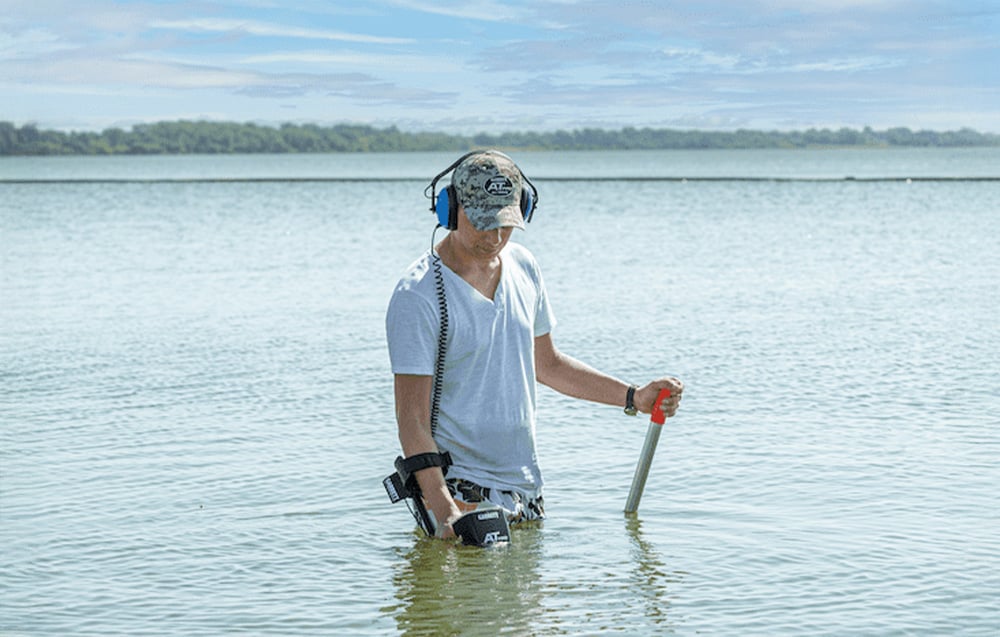 A man in the water uses a metal detector and holds a digging probe.