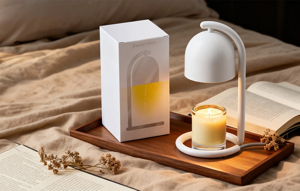 A white candle warmer standing on a wooden tray next to a white packaging box, with a small scented glass candle under the shade; in the background, an open boo