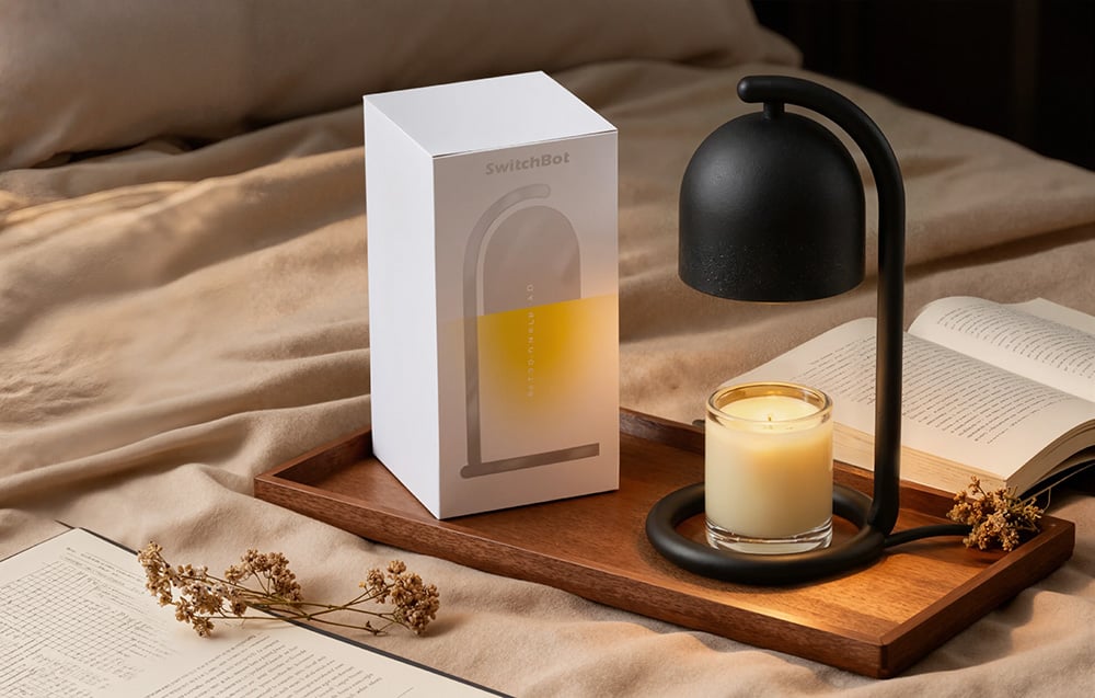 A black candle warmer standing on a wooden tray next to a white packaging box, with a small scented glass candle under the shade; in the background, an open boo