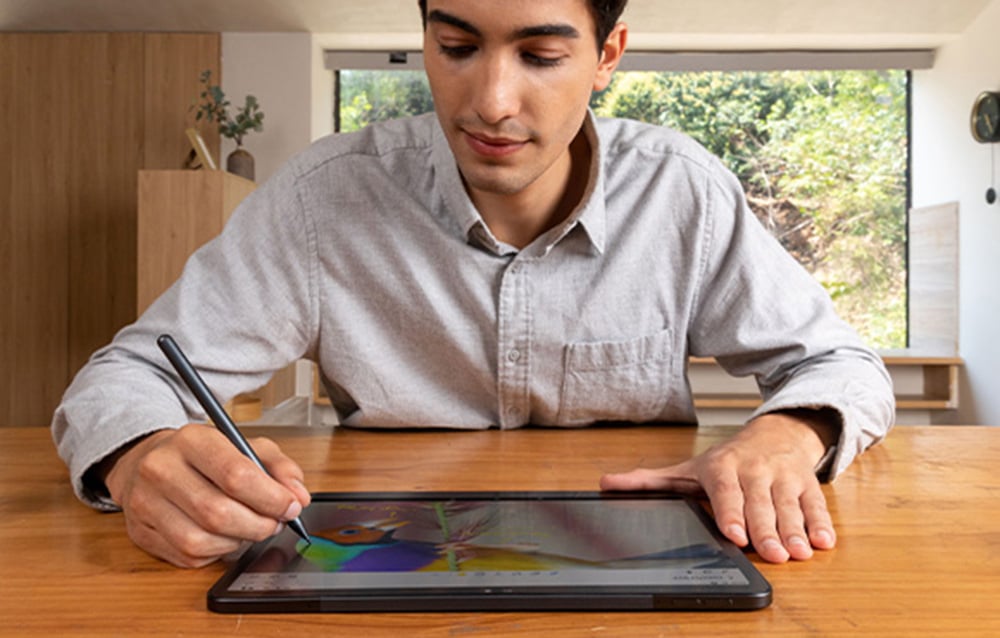 A man drawing with a stylus on a tablet lying flat on a wooden table at home