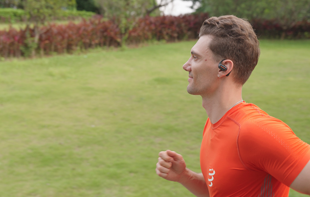 Man in orange T-shirt running on grass, wearing black open-ear headphones