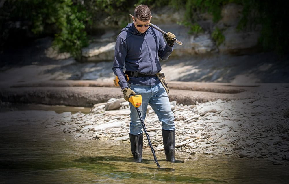 A man with a metal detector searches a shallow river in a wooded area.