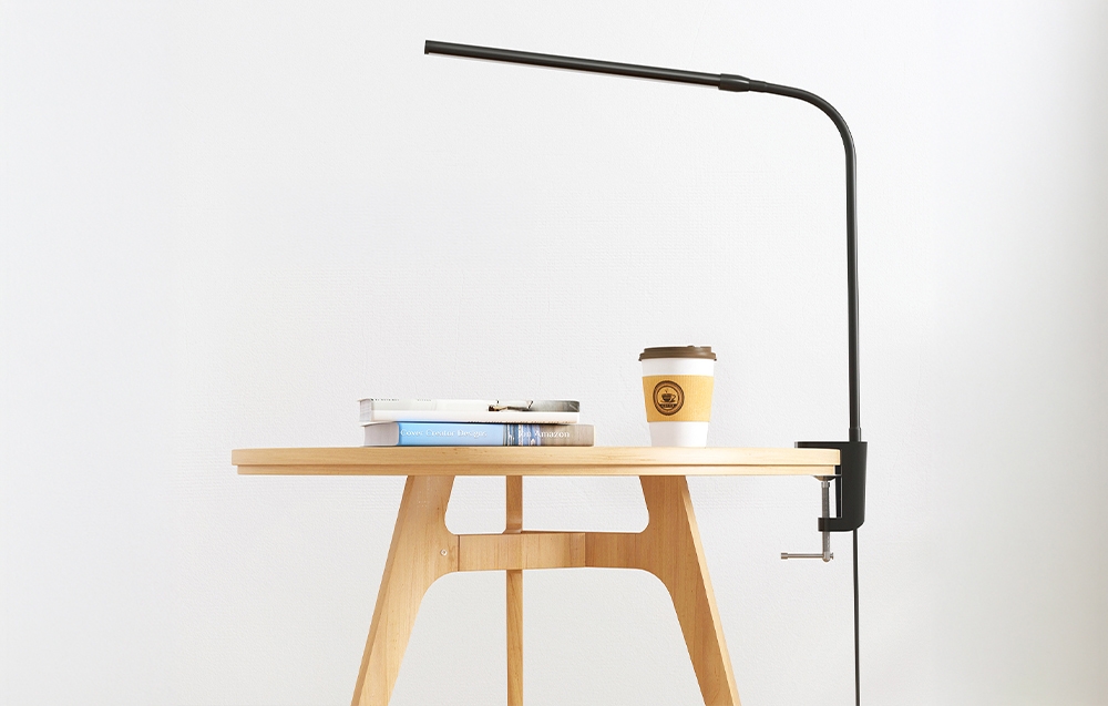 A flexible lamp attached to the edge of a round table. Next to it stands a stack of books and a paper cup of coffee.
