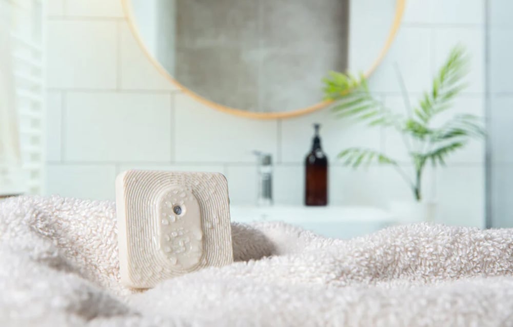 A beige Shelly H&T; Zigbee sensor with water droplets, lying on a fluffy white towel in a bright bathroom.