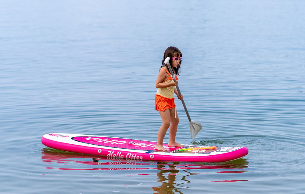 A girl wearing sunglasses stands on a white and pink swimming board with a paddle in her hand
