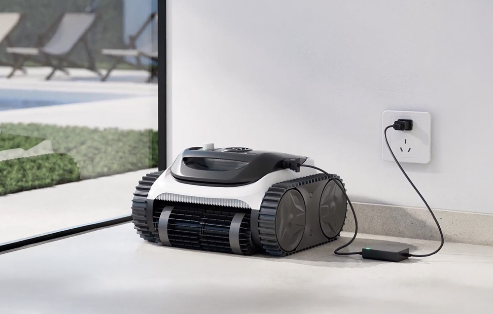 The pool robot stands on the terrace, plugged into a charger in a wall outlet next to a glass wall